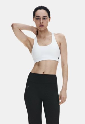 Femme aux cheveux foncés portant un soutien-gorge de sport blanc et un legging noir, posant avec une main derrière la nuque sur un fond uni.
