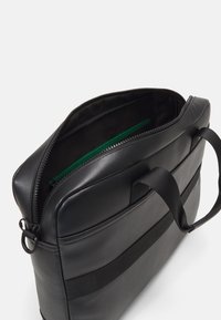 Borsa per laptop nera in materiale sintetico con apertura superiore con zip, caratterizzata da una texture liscia, una tasca interna verde e manici in tessuto regolabili.