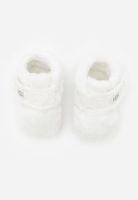 Białe, puszyste kapcie wykonane z miękkiego materiału, z paseczkiem wokół kostki i małą metką z napisem "UGG". Okrągły, przytulny kształt.