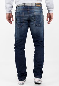 Jeans en denim bleu foncé à coupe droite, dotés de deux poches arrière et de coutures orange. Associés à des baskets blanches.