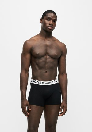 Atletyczny czarnoskóry mężczyzna stoi, ubrany w czarne bokserki z białym pasem i tekstem "JACK & JONES" na pasie, na jednolitym tle.