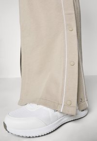 Beige pants with side snap buttons, white stitching, and a straight cut, paired with white textured athletic shoes.