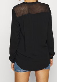 Chemise noire à manches longues en tissu doux, dotée d'un panneau arrière texturé semi-transparent et d'un ourlet arrondi. Associée à un short en denim.