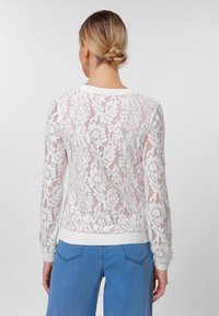 Λευκή δαντελένια πουλόβερ με λουλουδάτο σχέδιο, μακριά μανίκια και λευκό ριμπ κολάρο και μανσέτες. Φορίζεται με μπλε τζιν.