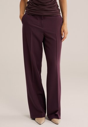 Femme portant un pantalon violet foncé taille haute à jambes larges avec poches, associé à des chaussures pointues beiges et un haut foncé froncé.