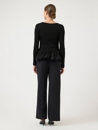 Sort langærmet top med flæsekant, kombineret med højtaljede, sorte bukser med brede ben og sorte hælede sko, vist bagfra.