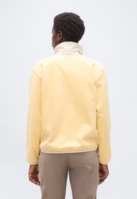 Άτομο με κοντά σγουρά μαλλιά που φοράει ένα ανοιχτό κίτρινο φλις μπουφάν και μπεζ παντελόνι, γυρισμένο πλάτη, με φόντο απλό.