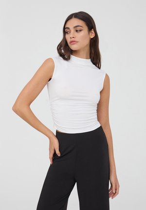 Top bianco senza maniche con collo alto e dettaglio arricciato ai lati, abbinato a pantaloni neri a vita alta. Tessuto di texture liscia.