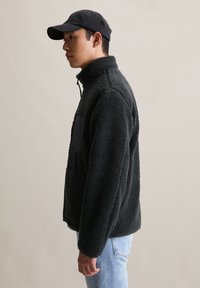 Homme en veste polaire noire et casquette se tenant de profil contre un fond uni clair.