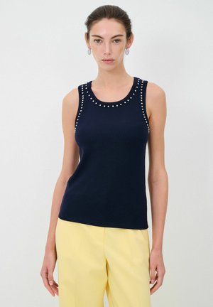 Top senza maniche di colore blu navy con scollatura rotonda, decorato con borchie argentate lungo le spalle, abbinato a pantaloni gialli a vita alta.