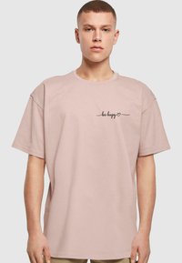 Lys rosa bomulds T-shirt med en afslappet pasform, rund halsudskæring, korte ærmer og sort kursiv tekst "være glad" på venstre bryst.