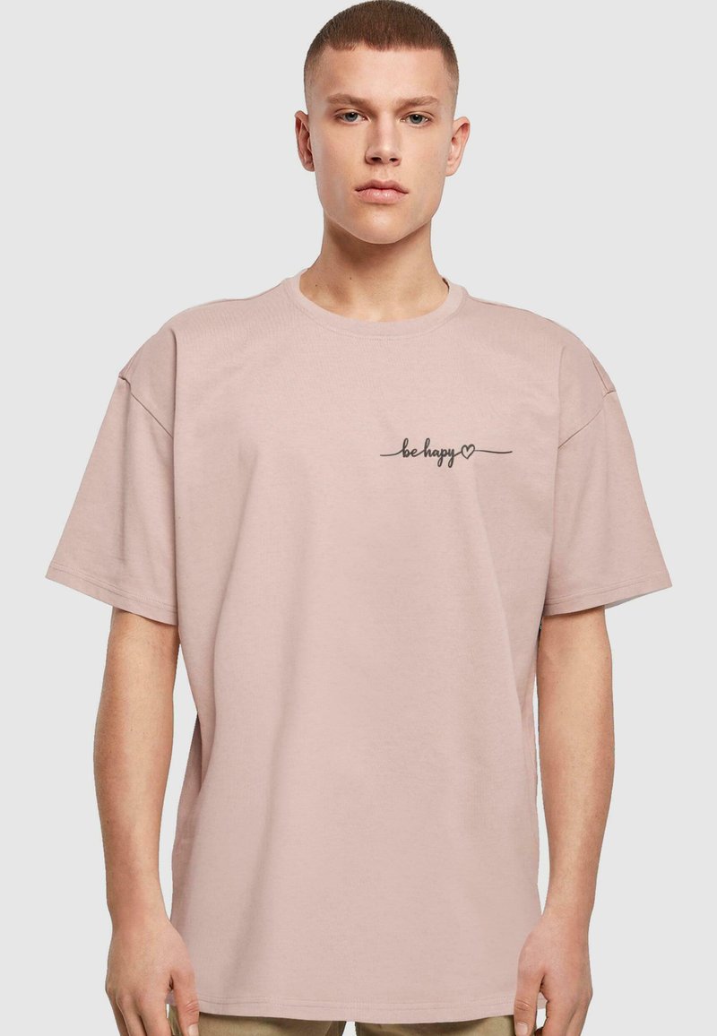 Lys rosa bomulds T-shirt med en afslappet pasform, rund halsudskæring, korte ærmer og sort kursiv tekst "være glad" på venstre bryst.