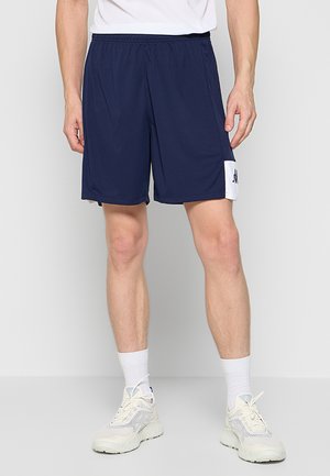 Uomo con pantaloncini sportivi blu navy con pannelli laterali bianchi, calzini bianchi e scarpe da corsa beige, in piedi su uno sfondo semplice.