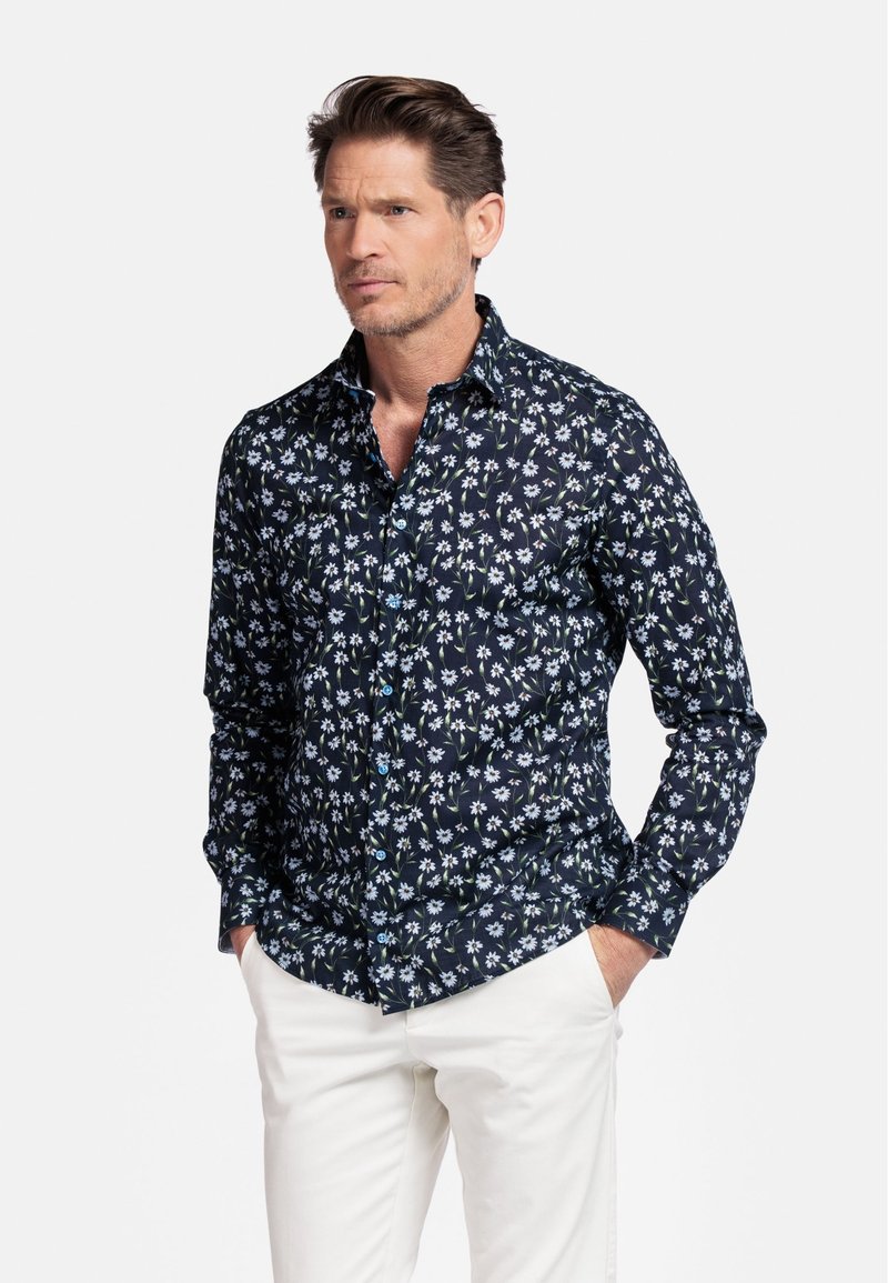 Chemise bleu marine avec motif floral blanc, manches longues, col boutonné et boutons bleus. Associée à un pantalon de couleur claire.