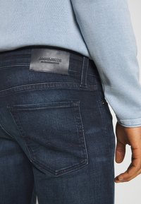 Mörka denimjeans med en läderlogga på midjebandet, synlig sömnad, fyra fickor och en slim fit-design.