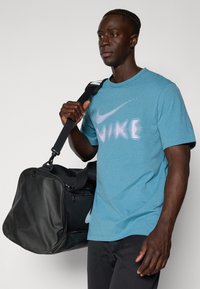Turkizna Nike majica s kratkimi rokavi, ki vsebuje gradientni logotip, v kombinaciji s črno športno torbo. Ima krojen ovratnik in gladko teksturo tkanine.