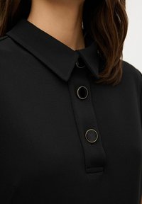 Camisa polo negra con cuello y tres botones, dos negros con detalles dorados y uno negro texturizado. Textura de tela suave.