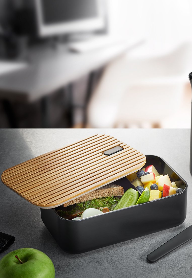 Schwarze rechteckige Bento-Box mit einem lamellenartigen Bambusdeckel. Enthält ein Sandwich, Gurkenscheiben und verschiedene Fruchtwürfel. Glatte Textur.
