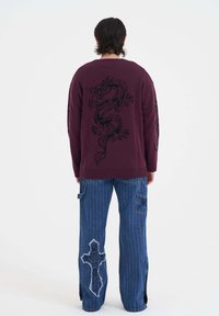 Bordový pletený svetr s černým designem draka na zadní straně. V kombinaci s modrými pruhovanými džíny, které mají křížovou nášivku a bočn í rozparky.