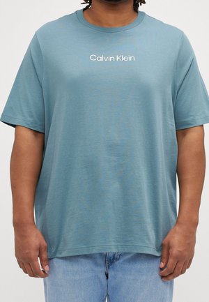 Person iført en teal Calvin Klein t-shirt med korte ærmer, kombineret med lyseblå denimjeans, vist fra nakke til hofter.