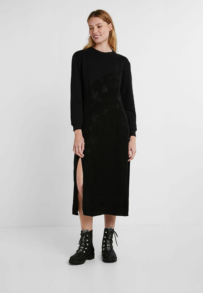 Zwarte jurk met lange mouwen, voorzien van een gestructureerd bloemenpatroon, een zijsplit en een ronde halslijn, gecombineerd met zwarte veterschoenen.