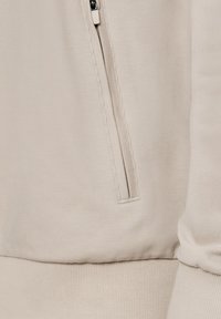 Beżowa tkanina z bliska, pokazująca kieszeń na zamek i ściągacz na mankiecie długiego rękawa.