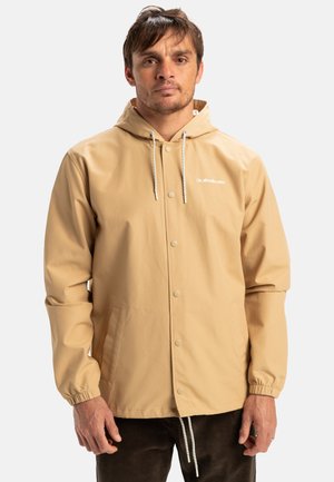 Beige waterdichte jas met een capuchon, voorzien van elastische manchetten, drukknoopsluiting en een merklogo op de borst. Glad textuur en lichtgewicht ontwerp.