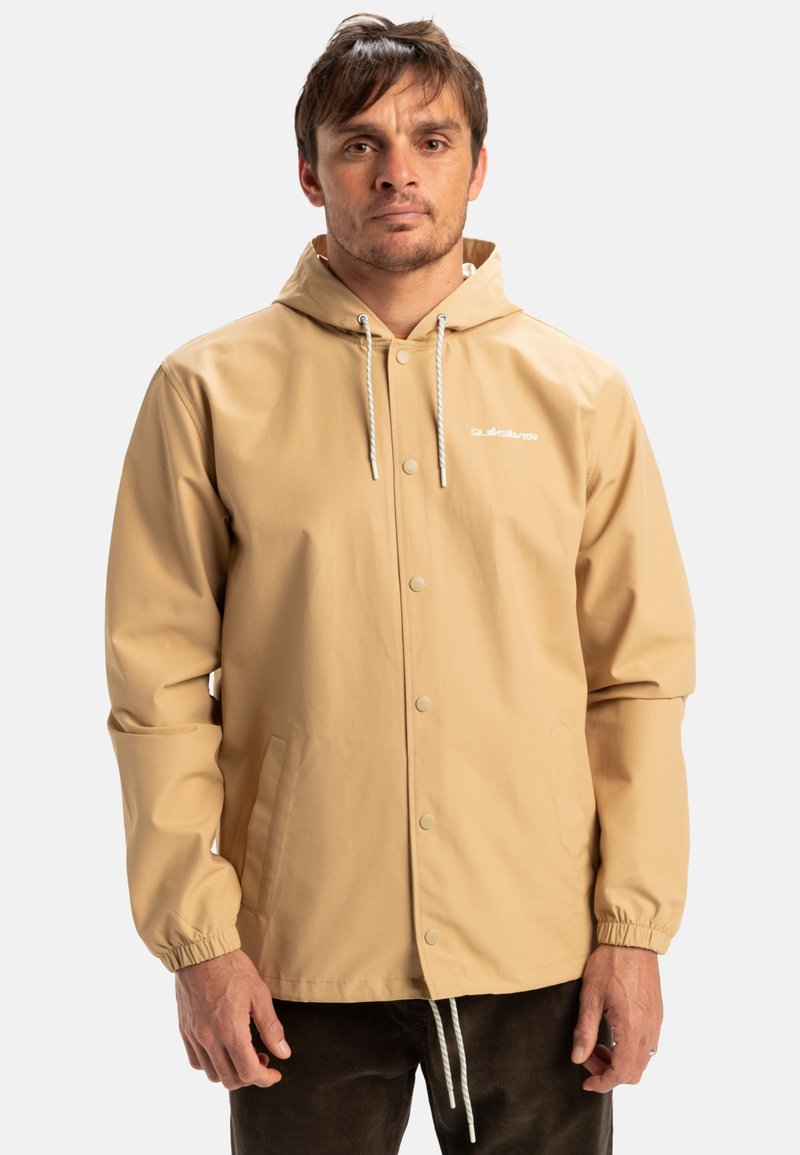 Beige waterdichte jas met een capuchon, voorzien van elastische manchetten, drukknoopsluiting en een merklogo op de borst. Glad textuur en lichtgewicht ontwerp.