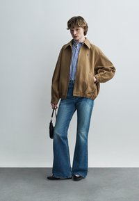 Persona che indossa una giacca marrone chiaro, camicia a righe blu e bianca, jeans a zampa, scarpe nere, che tiene una piccola borsa nera, in piedi davanti a uno sfondo chiaro e uniforme.