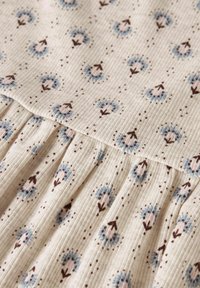 Tejido de color beis claro con una textura acanalada, que presenta un patrón floral con flores azules y rosas, acentuado por tallos marrones y puntos moteados.