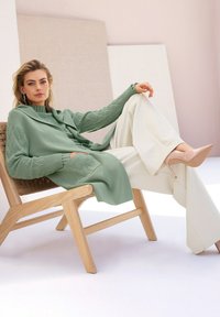 Vrouw die achterover leunt op een houten stoel, gekleed in een groene gebreide trui en sjaal, witte wijde broek en beige hoge hakken.