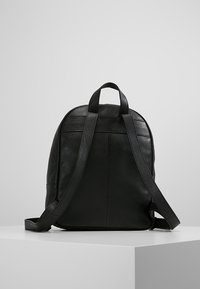 Svart läderryggsäck med två justerbara axelremmar och ett handtag på toppen, uppvisad på en vit pelare mot en grå bakgrund.