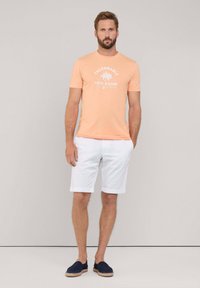 Camiseta melocotón con gráfico de palmera, pantalones cortos blancos y espadrilles azul marino. El conjunto presenta un diseño casual y ligero, adecuado para el clima cálido.