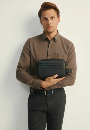 Czarna skórzana saszetka z zamkiem błyskawicznym, podwójnymi przegródkami i gładką fakturą, trzymana przez modela w brązowej koszuli zapinanej na guziki i ciemnych spodniach.