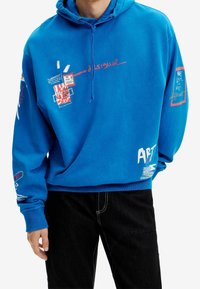 Modrá mikina s kapucí s grafickými potisky v červené a bílé barvě. Má kapuci se stahovací šňůrkou, žebrované manžety a různé textové a vzorové detaily na rukávech.