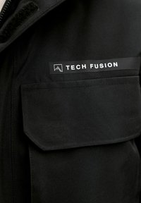 Sort jakke med en brystlomme, der har en rektangulær label med teksten "TECH FUSION" i hvidt; glat stof og minimalistisk design.