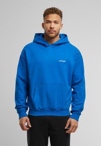 Sudadera con capucha azul, hecha de algodón, con un bolsillo frontal, puños acanalados y un pequeño logo en el pecho. Textura suave, corte relajado.