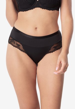 Kobieta ubrana w czarne majtki z wysokim stanem ozdobione koronką oraz pasujący czarny koronkowy biustonosz, stojąca na tle białego tła.
