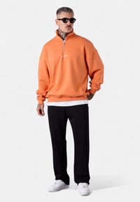 Orangener Sweatshirt mit Viertelreißverschluss-Kragen, weißem Logo auf der Brust, schwarzen Hosen und weißen Sneakers. Einfarbig, lockere Passform.