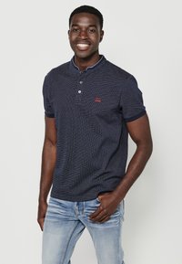 Jūrininkų mėlynos spalvos trumpomis rankovėmis polo marškiniai su smulkia rašto tekstūra ir kontrastinėmis apykaklėmis, krūtinėje įspaustas raudonas logotipas. Apačioje matomi džinsai.