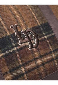 Ruda langinė vilnos medžiaga su tamsiu išsiuvinėtu logotipu. Dizainas apima žalios ir smėlio spalvos atspalvius languotėje.