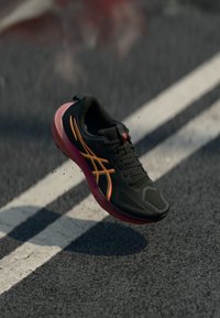 Chaussure de course noire avec une tige texturée, des accents orange et jaune, et une semelle rose, montrée en l'air au-dessus d'une piste grise.