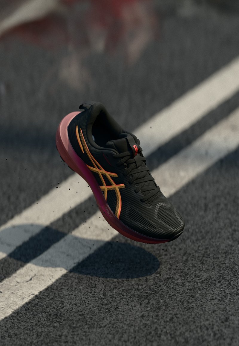 Chaussure de course noire avec une tige texturée, des accents orange et jaune, et une semelle rose, montrée en l'air au-dessus d'une piste grise.