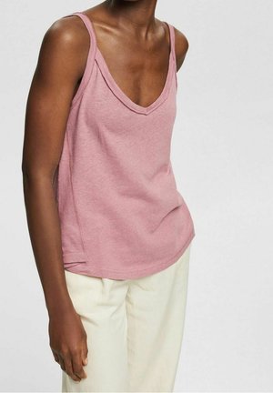 Persona di colore che indossa un top rosa senza maniche con scollo a V e pantaloni beige chiaro, mostrata dalla spalla a metà coscia su uno sfondo semplice.