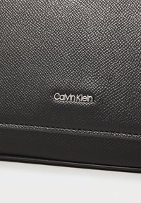 Portafoglio in pelle nera testurizzata con logo Calvin Klein argentato. Le caratteristiche includono cuciture definite e un design moderno e liscio.