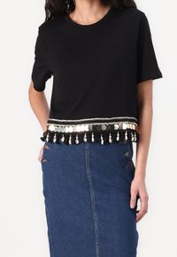 Camiseta negra recortada de manga corta, con un ribete decorativo de monedas y borlas en el dobladillo. Combinada con una falda de mezclilla.