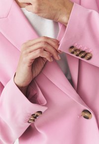 Blazer rosa con rever strutturati, tessuto texturizzato, quattro bottoni marroni sui polsini e accenti di cucitura rosa. Top bianco visibile sotto.