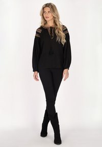 Schwarze Bluse mit gestickten Schulterakzenten, mit einer Schleifenmasche am Hals und Puffärmeln, kombiniert mit schwarzen engen Hosen und schwarzen kniehohen Stiefeln.