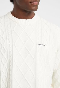 Maglione bianco a maglia lavorata a treccia con un motivo a diamante, scollo a costine e una piccola etichetta "GUESS JEANS" sulla parte anteriore.