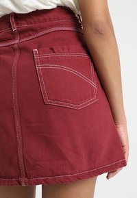 Jupe en denim bordeaux avec une taille ajustée, des coutures blanches visibles et une poche arrière au design incurvé. Texture douce, longueur courte.